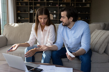Worried concerned young couple doing paperwork at home, using laptop computer, talking, discussing...