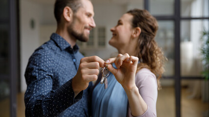 Happy cheerful young couple moving into new home, holding showing key. Married homeowners, tenant having fun at housewarming party, dancing, hugging, smiling, talking. Real estate property buying
