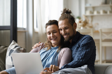 Cheerful happy family couple watching movie on laptop in living room, resting on comfortable couch with computer, using online app, internet service, laughing, talking on video call together