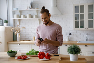 Happy cheerful millennial guy cooking salad in home kitchen, consulting recipe on internet, chatting online, watching food blog, making call on mobile phone at table with fresh vegetables ingredients