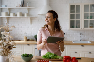 Happy millennial girl enjoying cooking salad from fresh vegetables in kitchen, using digital tablet pad, consulting recipe, healthy food blog, thinking over dinned menu, looking away, smiling