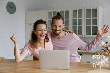 Cheerful excited couple celebrating achieve at computer, receiving good news on video call, email, online notification, message on internet, hugging at laptop in kitchen, feeling happy, laughing