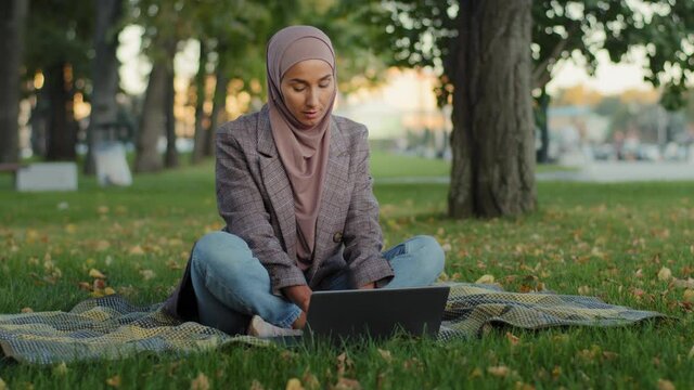 Muslim arab islamic student girl in hijab business woman freelancer user uses laptop sitting on green grass in park working studying remotely in summer quarantine chatting browsing online shopping