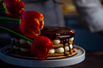 A cake with honey cakes and white cream stands on a white stand and is poured with chocolate on top, next to it are tulips, all in warm colors. High quality photo