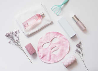 Facial cosmetic with pink sheet mask, falcons with empty label mock up, candles, massage tools and flowers on white background. Themed beauty concept. Top view with copy space.