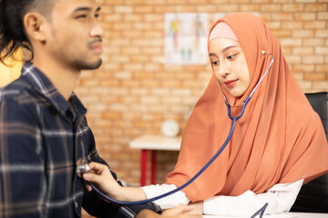 Young beautiful woman doctor is health examining male patient in office of hospital clinic and advising with a smile on medicines. This Asian medical specialist is an Islamic person wearing a hijab.