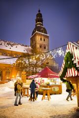 Fototapeta premium Winter evening on the main square of the city, street by evening lights, before Christmas, New Year. Christmas Fair. Catholic cathedral, church, tower. Riga, Latvia.