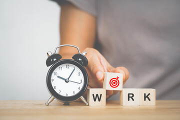 senior's hand holding dartboard icon on wood cube, part of work word, and black analog alarm clock...
