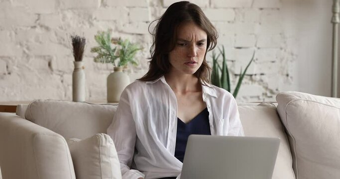 Young 20s woman sit on sofa with laptop, typing text, working or studying online, feels angry, irritated and concerned by mistake in task, computer malfunction, incorrect or slow device work concept
