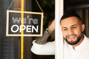 Smiling young Latin American man at the entrance of a small business next to a sign with the text We're Open.