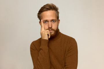 Horizontal picture of sad bored male in warm brown wool sweater, holding fingers on temple, looking at camera with tired, upset face expression. Human emotions, feelings, body language, gestures