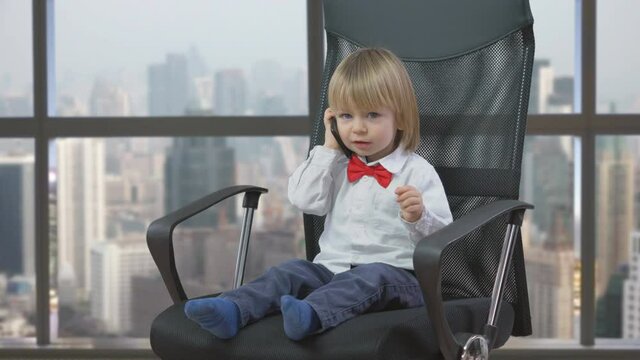 Portrait of little lovely blonde hair child with red bow sit on business chair and talk on phone, window office city view