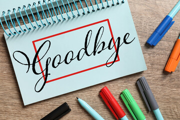 Notebook with word Goodbye and markers on wooden table, flat lay