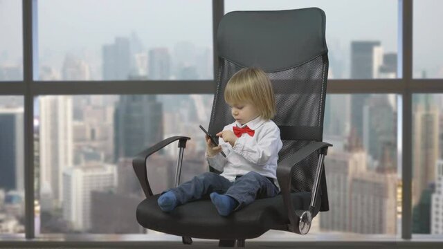 Little child with red bow sit on big chair and searching on mobile phone, modern entertainment, harmful habit, office window city panorama