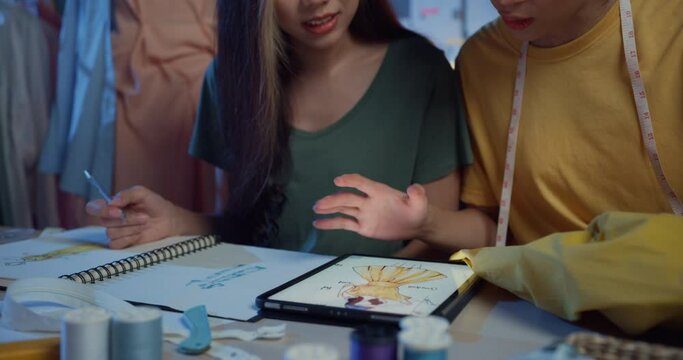 Close-up two professional Asia cheerful lady fashion designer teamwork with casual sitting on table use tablet meeting discuss final design share idea in shop at night. Lady tailor start up business.