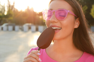 Beautiful young woman eating ice cream glazed in chocolate on city street, closeup