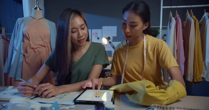 Two professional Asia cheerful lady fashion designer teamwork with casual sitting on table use tablet meeting discuss fabric final design share idea in shop at night. Lady tailor start up business.