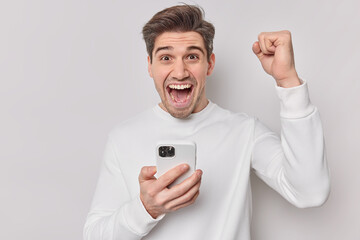 Photo of joyful adult man clenches fists and holds mobile phone celebrates success rejoices good...