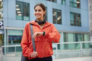 Waist up shot of happy brunette sporty woman dressed in anorak checks fitness results on smartwatch carries fitness mat waits for someone poses against modern city building. Healthy lifestyle