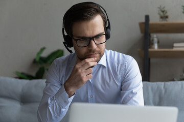 Serious businessman wear headset looks at laptop working at home, thinking over task, develop online project, listening audio course improve foreign language skills. E-learning use modern tech concept