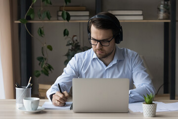 Millennial serious concentrated man sit at workplace desk holds pen noting information, makes assignment listen audio course through wireless headset. Improve foreign language skills, e-study concept