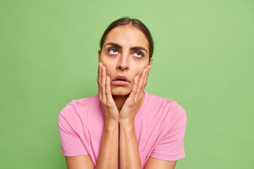 Uninterested bored young woman keeps hands on cheeks fed up of dull conversation looks above makes...