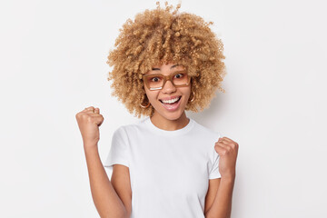 Happy triumphing young woman with curly hair clenches fists celebrates success glad to achieve...
