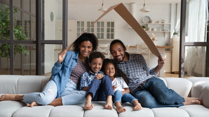 Beautiful African couple with children sitting at home on sofa under symbolic cardboard roof...