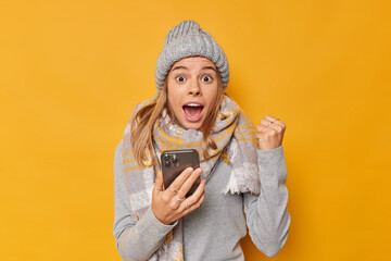 Sweet taste of success. Positive excited woman gained her daily app in goal holds mobile phone clenches fist exclaims loudly wears knitted hat jumper and scarf around neck isolated over yellow wall