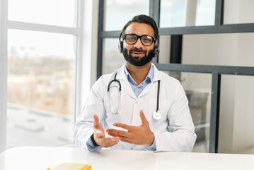 Confident Indian male MD wearing headset, eyeglasses and medical gown giving online consultation,...
