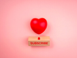 Subscribe concept. Word "SUBSCRIBE" and bell icon on wooden cube block and red heart ball on pink pastel background, minimal style. Following social media.