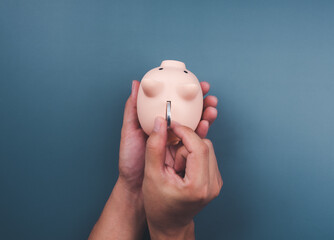 Pink piggy bank on a blue background while hand holding it and putting a silver coin, top view, minimal style. Saving money and business investment concept. © tete_escape