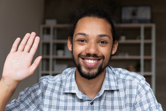 Close up smiling African guy sit indoor looking at camera raise palm wave hand start video call remote communication, webcam view. Video conference participant profile picture, virtual meeting concept