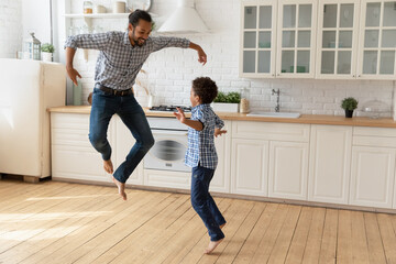 African father and little adorable son jumping have fun in modern kitchen, listen music dance...