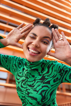 Vertical shot of happy playful woman with bun hairstyle keeps palms near face winks eye and smiles broadly has vivid makeup dressed in green jumper expresses positive emotions foolishes around in city