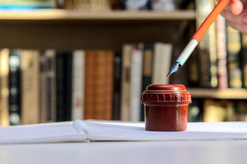 In a woman's hand is a fountain pen, which she dips into an inkwell. Background - books.