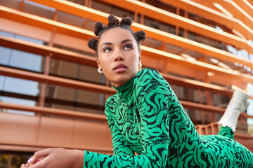 Horizontal shot of pretty young woman with hair buns applies blue eyeliner dressed in green costume and boots focused into distance during daytime considers something. People fashion style concept - obrazy, fototapety, plakaty