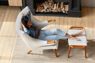 Top view peaceful African woman relax on armchair in modern cottage house with fireplace, put feet...