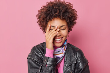 Fototapeta na wymiar Indoor shot of happy curly haired young woman keeps eyes closed hand on face smiles gently wears leather jacket isolated over pink studio bacground. Positive emotions and sincere feelings concept