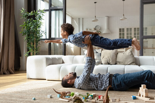 African dad lying on floor raised cute 5s little son on outstretched arms, boy imagines flying in air, pretend superhero, having fun together at warm home. Dreams about travel, family vacation concept