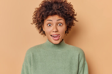 Surprised happy woman with curly hair licks lips with tongue looks with curious expression at something interesting dressed in casual jumper isolated over beige background. Human reactions concept