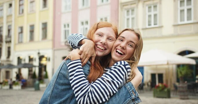 Two young women girls hugging, cuddling, looking to camera on street in city. Pretty Caucasian female best friends snuggling and laughing. Friendship, relationship, feminity concept.