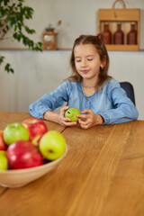 Fototapeta premium Happy little girl takes a pear from the bowl with variety fruits on table at home