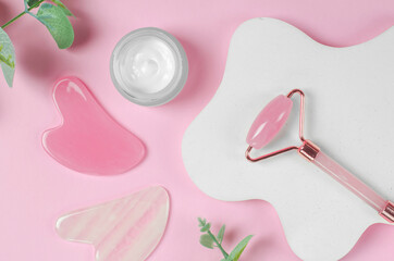 Roller jade facial massager and open jar of moisturizer on pink background with geometric shapes. Gua Sha face massage tools on pink background with green leaves