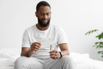 Serious young black bearded male sitting on bed in bedroom, hold glass of water and tablet