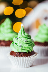 Chocolate cupcakes or muffins decorated as Christmas tree with buttercream. Ginger house and Christmas lights from Christmas tree in blurry background. Festive and holiday decoration