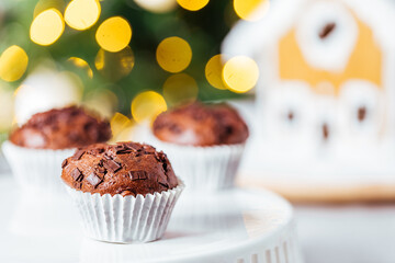 Chocolate cupcakes or muffins with Ginger house and Christmas lights from Christmas tree in blurry background. Festive and holiday decoration
