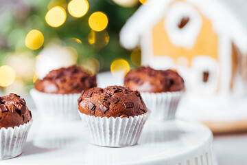 Chocolate cupcakes or muffins with Ginger house and Christmas lights from Christmas tree in blurry background. Festive and holiday decoration