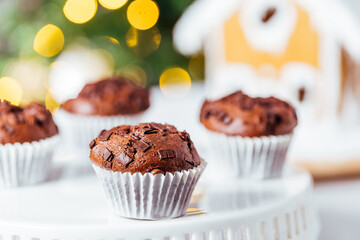 Chocolate cupcakes or muffins with Ginger house and Christmas lights from Christmas tree in blurry background. Festive and holiday decoration