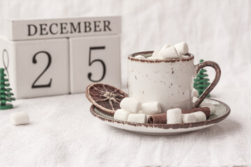 Hot cocoa or coffee with marshmallow in a white ceramic mug, White block calendar present date 25 December on a white background. The concept of Christmas morning, cozy holidays and New Year